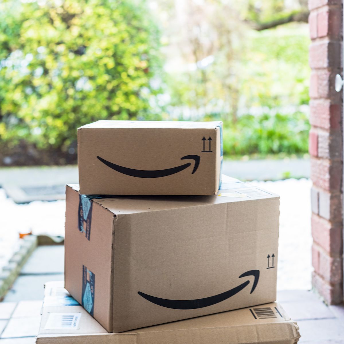 Two brown Amazon boxes with the smile logo sit on a front porch. Amazon brushing scams happen when retailers send you unordered packages in hopes to use your info to boost their sales.
