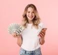 Young blond woman smiling holding cash in one hand and cellphone on the other