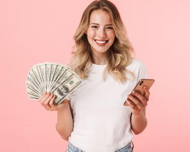 Young blond woman smiling holding cash in one hand and cellphone on the other