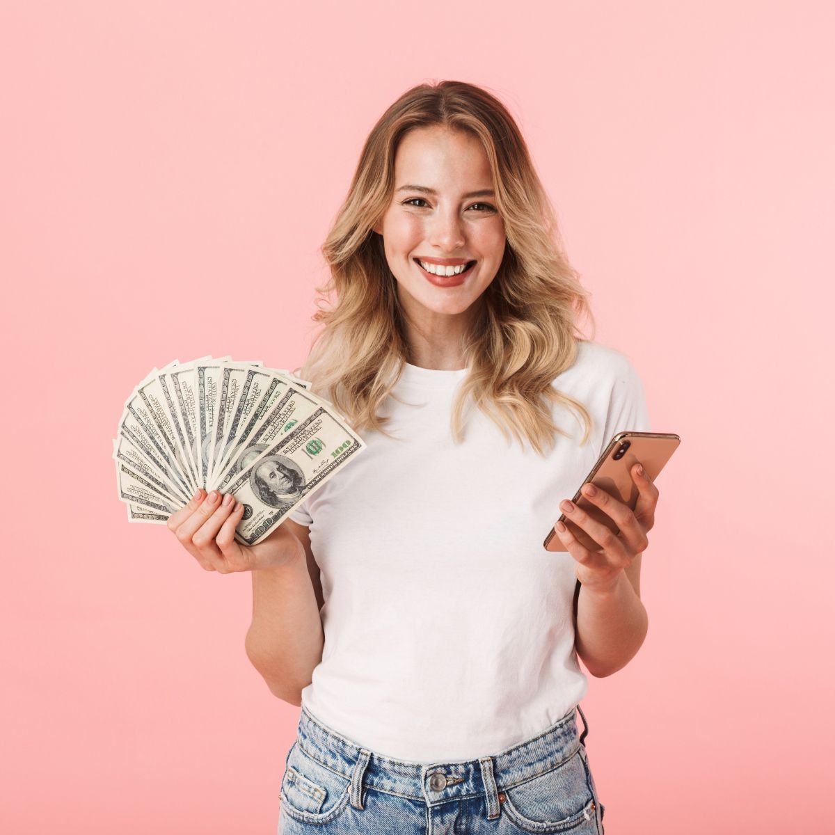 Young blond woman smiling holding cash in one hand and cellphone on the other