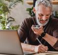 A senior man writes on a notepad while talking on speakerphone and sitting in front of a laptop