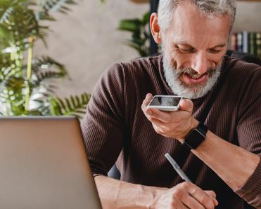 A senior man writes on a notepad while talking on speakerphone and sitting in front of a laptop