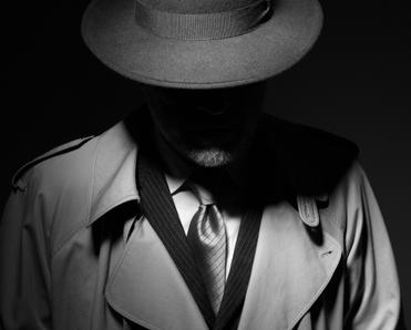 A black and white, moody photo of a man dressed like an old school private investigator with a hat and trench coat.