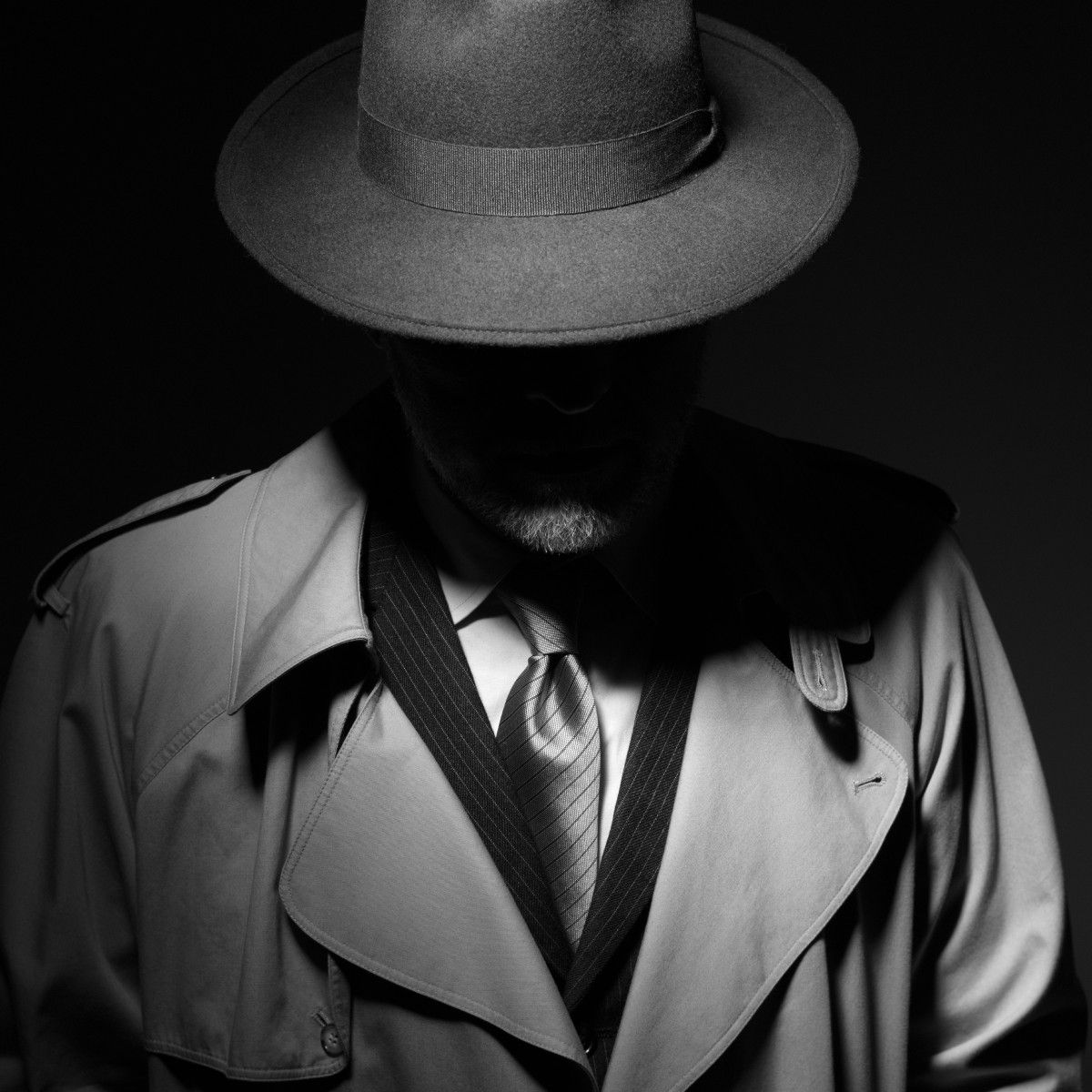 A black and white, moody photo of a man dressed like an old school private investigator with a hat and trench coat.