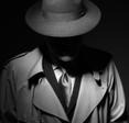 A black and white, moody photo of a man dressed like an old school private investigator with a hat and trench coat.