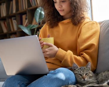 A young Hispanic woman sits on a gray couch next to a tabby cat. She&#x27;s holding a yellow mug and her laptop is in her lap to illustrate having a good internet download and upload speed.