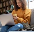 A young Hispanic woman sits on a gray couch next to a tabby cat. She&#x27;s holding a yellow mug and her laptop is in her lap to illustrate having a good internet download and upload speed.