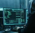 Computer screen with green neon coding language and a hooded hacker in front typing on keyboard