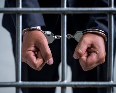A close-up of a man&#x27;s hands in handcuffs as he&#x27;s behind a jail cell.