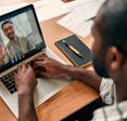 A rear-view of a Black man video conferencing with his coworker on Zoom. The desk is covered with paperwork, and the coworker on the screen is waving and smiling.