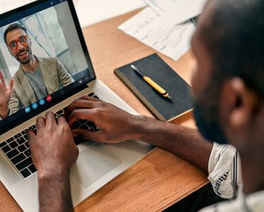 A rear-view of a Black man video conferencing with his coworker on Zoom. The desk is covered with paperwork, and the coworker on the screen is waving and smiling.