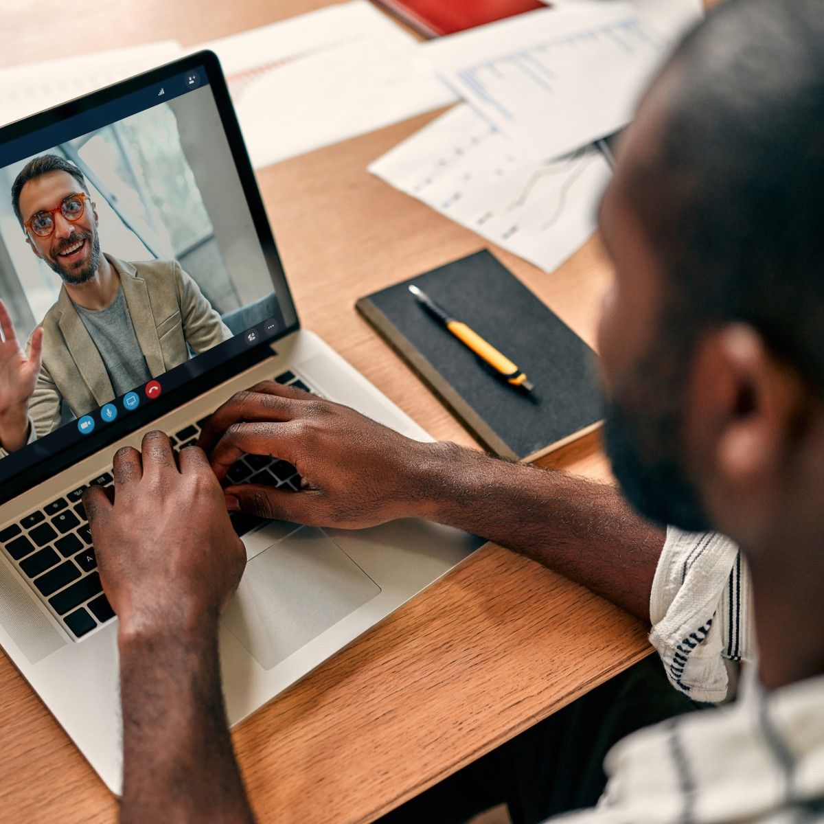 A rear-view of a Black man video conferencing with his coworker on Zoom. The desk is covered with paperwork, and the coworker on the screen is waving and smiling.