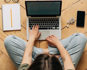 A top-down view of a dark-haired woman wearing jeans and green shirt typing on her Macbook laptop. She&#x27;s sitting on a wooden floor and her cell phone, earbuds, and a notebook and pencil are surrounding her.