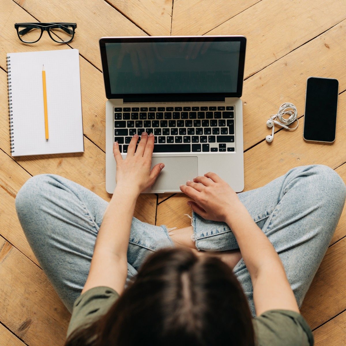 A top-down view of a dark-haired woman wearing jeans and green shirt typing on her Macbook laptop. She&#x27;s sitting on a wooden floor and her cell phone, earbuds, and a notebook and pencil are surrounding her.