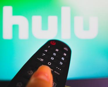 A close-up photo of a woman&#x27;s hand holding a remote and pointing it at her TV. On the screen is the Hulu logo.