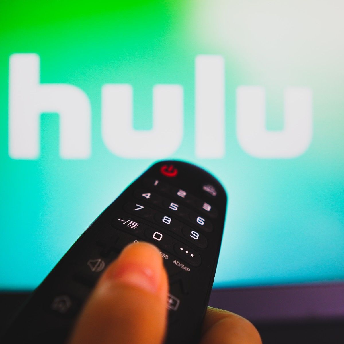 A close-up photo of a woman&#x27;s hand holding a remote and pointing it at her TV. On the screen is the Hulu logo.