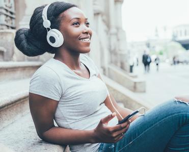 A young Black woman sits on steps outside and listens to music on Spotify.