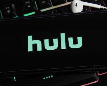 A close-up of a smartphone screen with the Hulu logo in green on a black background. The phone is sitting on top of a gaming computer keyboard.