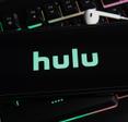 A close-up of a smartphone screen with the Hulu logo in green on a black background. The phone is sitting on top of a gaming computer keyboard.