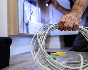 An internet technician holds a coil of Ethernet cable while he shines a flashlight on a wall outlet.