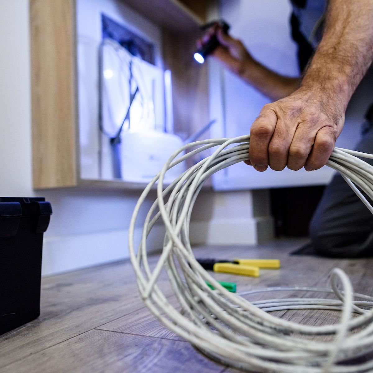 An internet technician holds a coil of Ethernet cable while he shines a flashlight on a wall outlet.