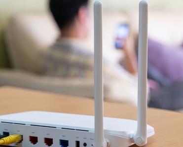 A white Wi-Fi router sits on top of a light-grained wood cabinet. In the background, a man sits on a white sofa and looks at his cell phone.