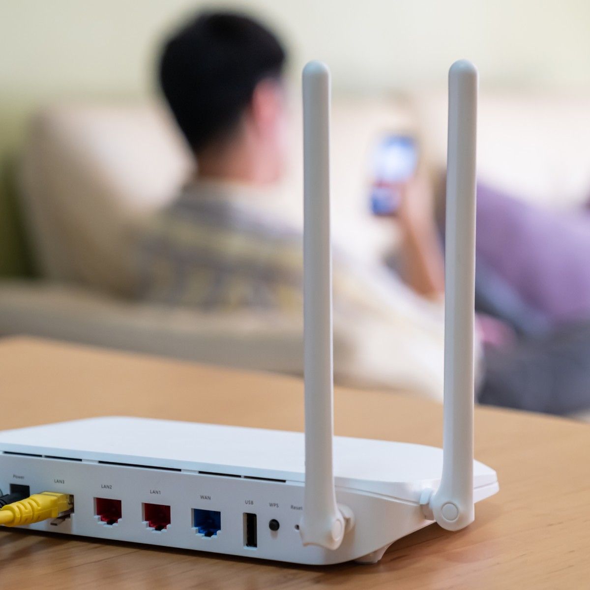 A white Wi-Fi router sits on top of a light-grained wood cabinet. In the background, a man sits on a white sofa and looks at his cell phone.