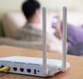 A white Wi-Fi router sits on top of a light-grained wood cabinet. In the background, a man sits on a white sofa and looks at his cell phone.