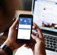 A man holds his cell phone in front of him, the Facebook login screen is displayed. In the background, his Facebook news feed is pulled up on his laptop.