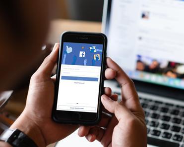 A man holds his cell phone in front of him, the Facebook login screen is displayed. In the background, his Facebook news feed is pulled up on his laptop.