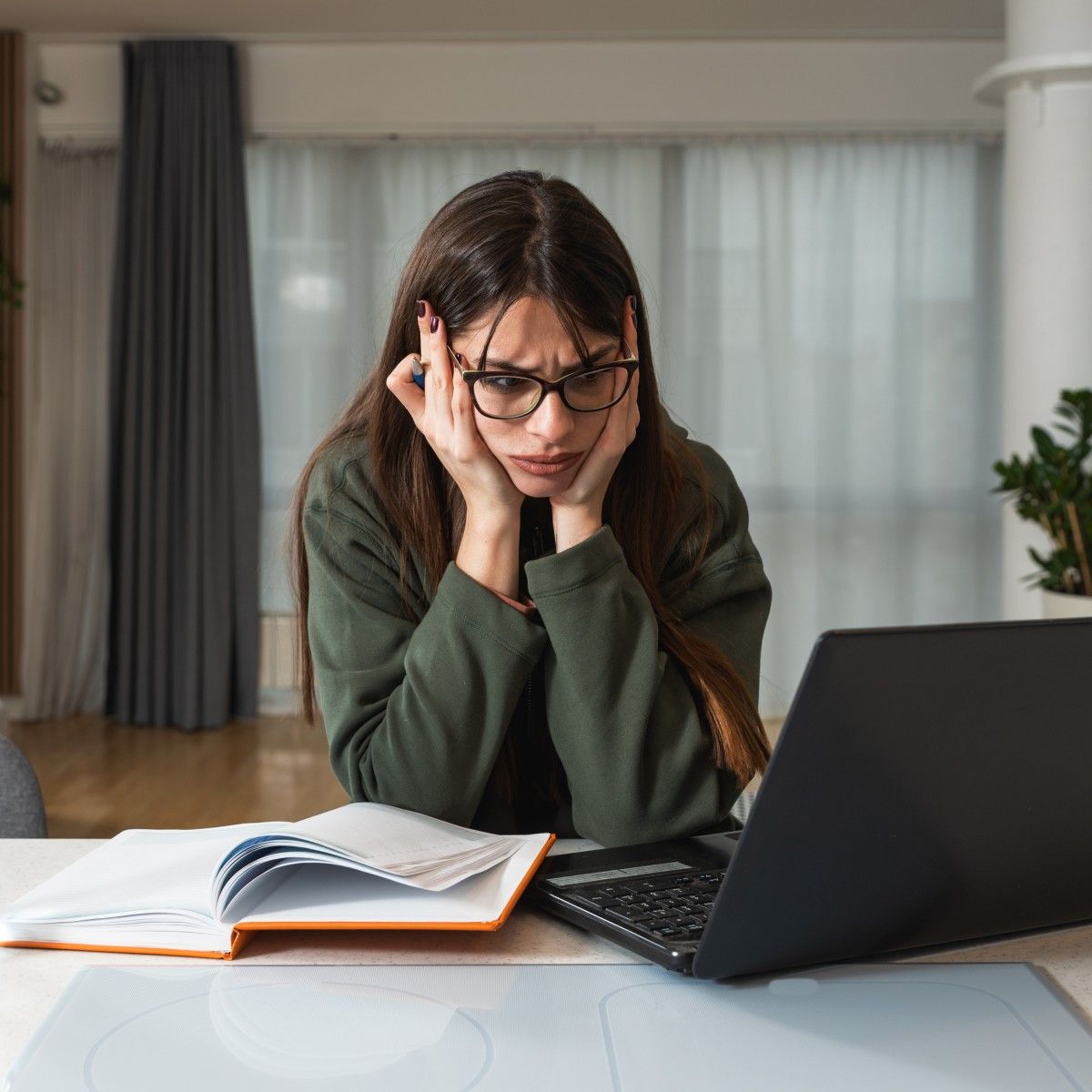 A young, dark-haired woman with glasses and a forest green sweatshirt holds her head in her hands and looks in frustration at her laptop.