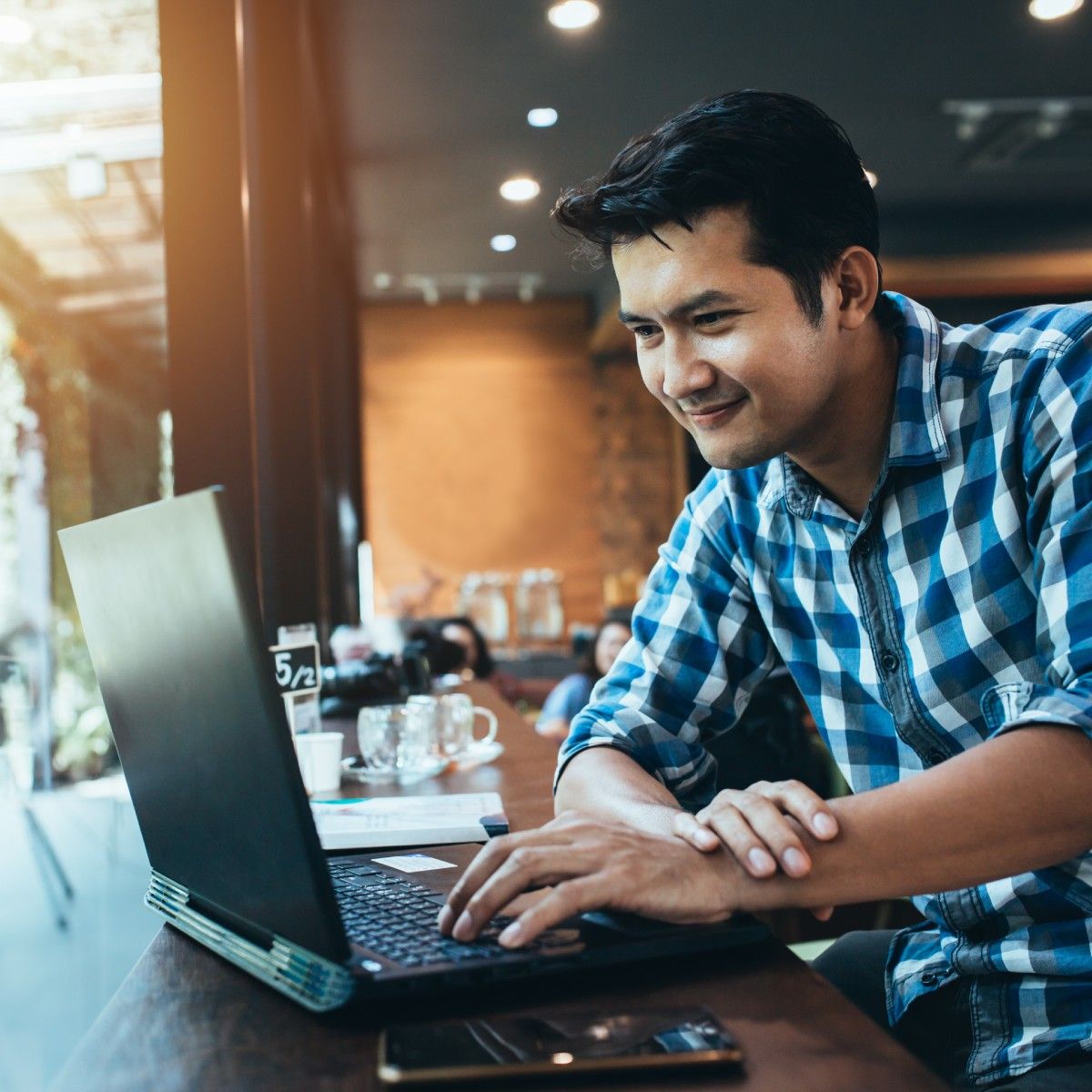 An Asian man in a blue plaid shirt types on his laptop while sitting next to a window in a coffee shop.