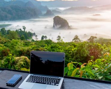 A laptop and cell phone sit on a table facing a gorgeous view of the sun rising over a jungle and mountains.