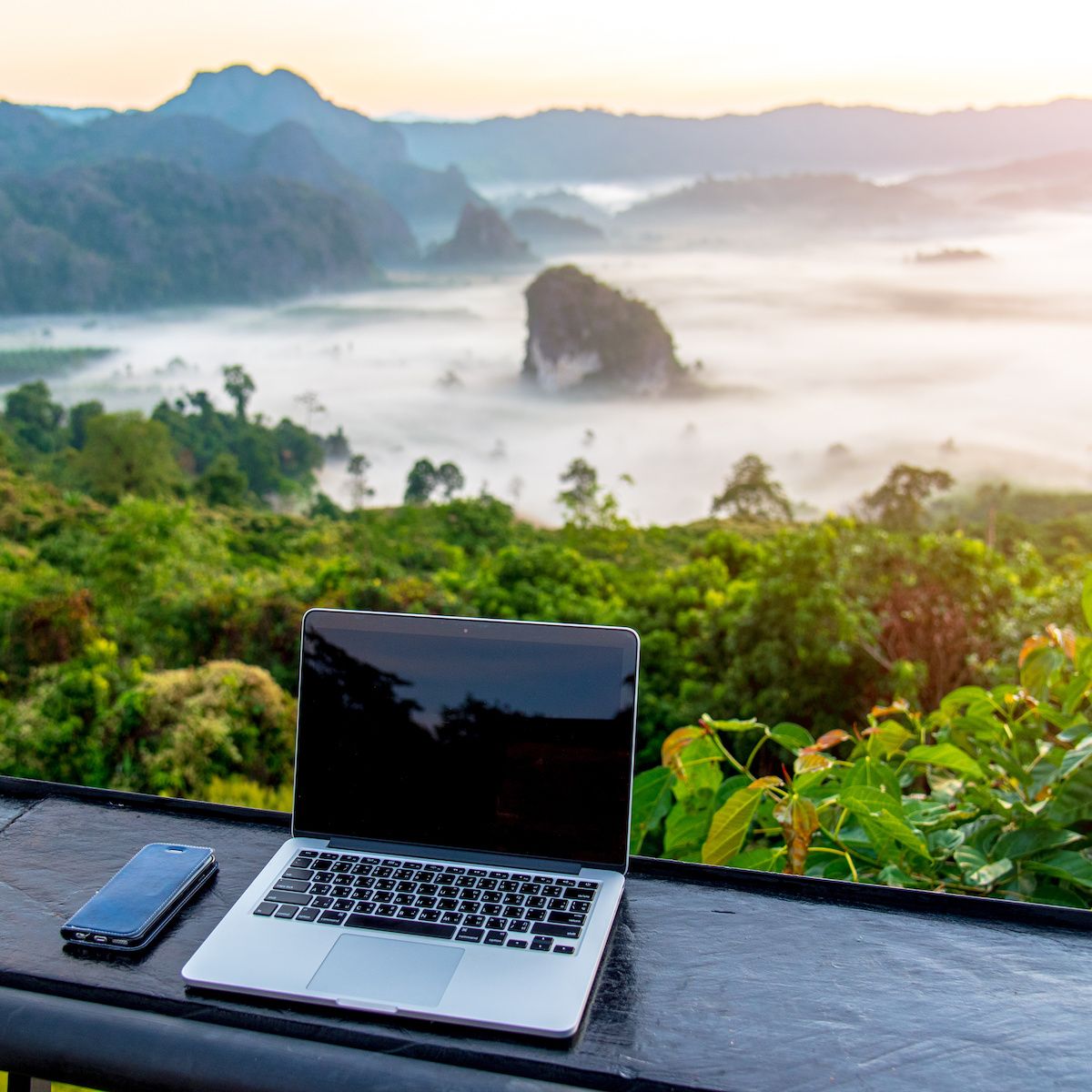 A laptop and cell phone sit on a table facing a gorgeous view of the sun rising over a jungle and mountains.