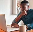 Man thinking, typing, and working on a laptop at his desk with a coffee mug by his side.