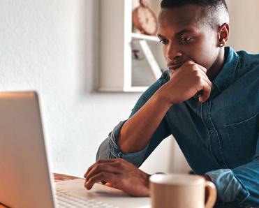 Man thinking, typing, and working on a laptop at his desk with a coffee mug by his side.