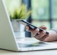 A close-up of a hand using an iPhone while hovering above a laptop.