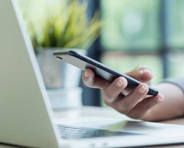 A close-up of a hand using an iPhone while hovering above a laptop.