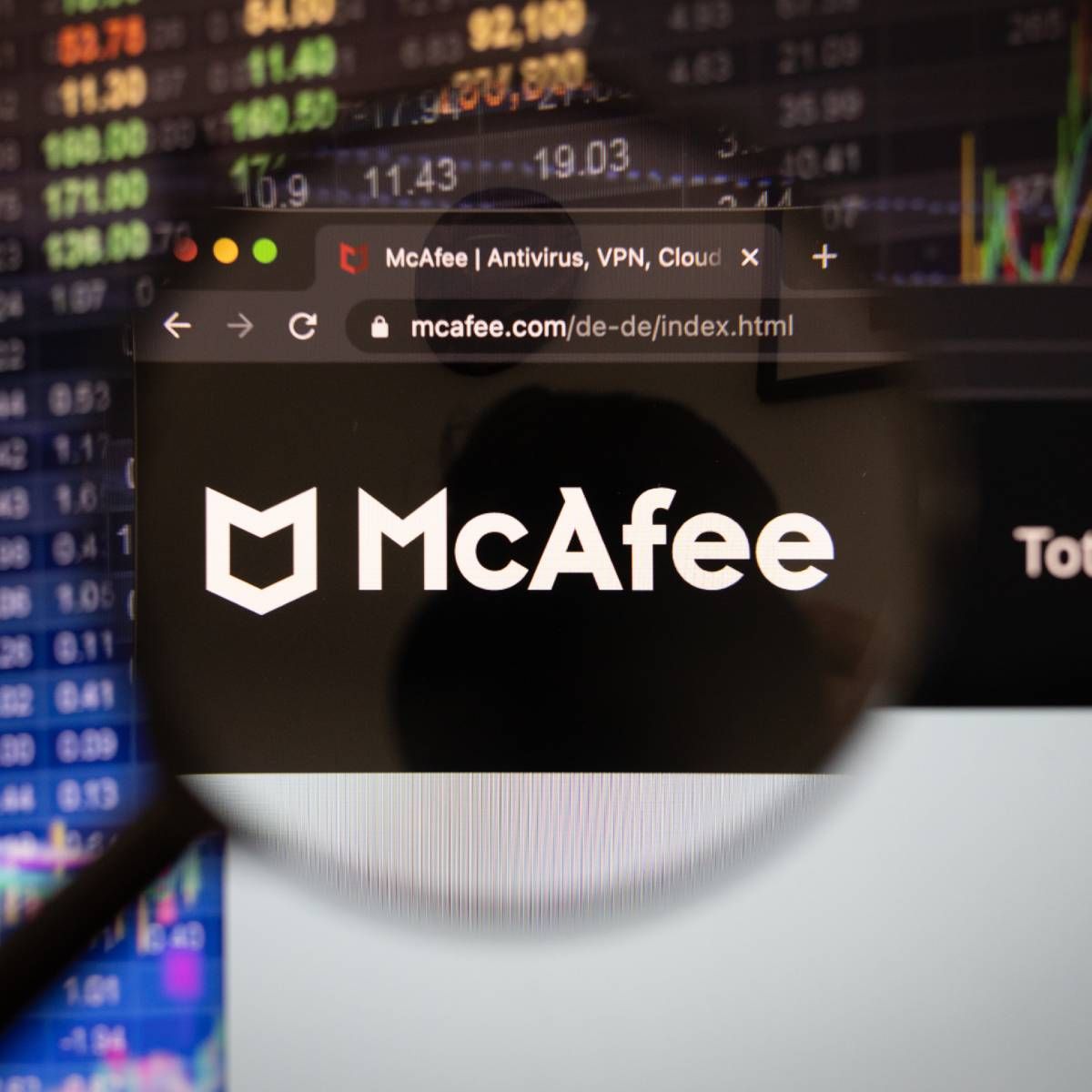 McAfee company logo on a website with blurry stock market developments in the background, seen on a computer screen through a magnifying glass.