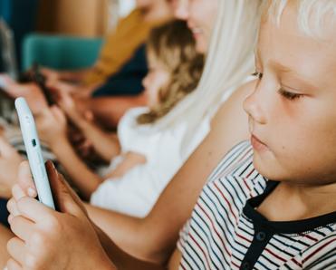 A young boy with blond hair sits on the couch with his siblings and uses internet data while playing on a cell phone.