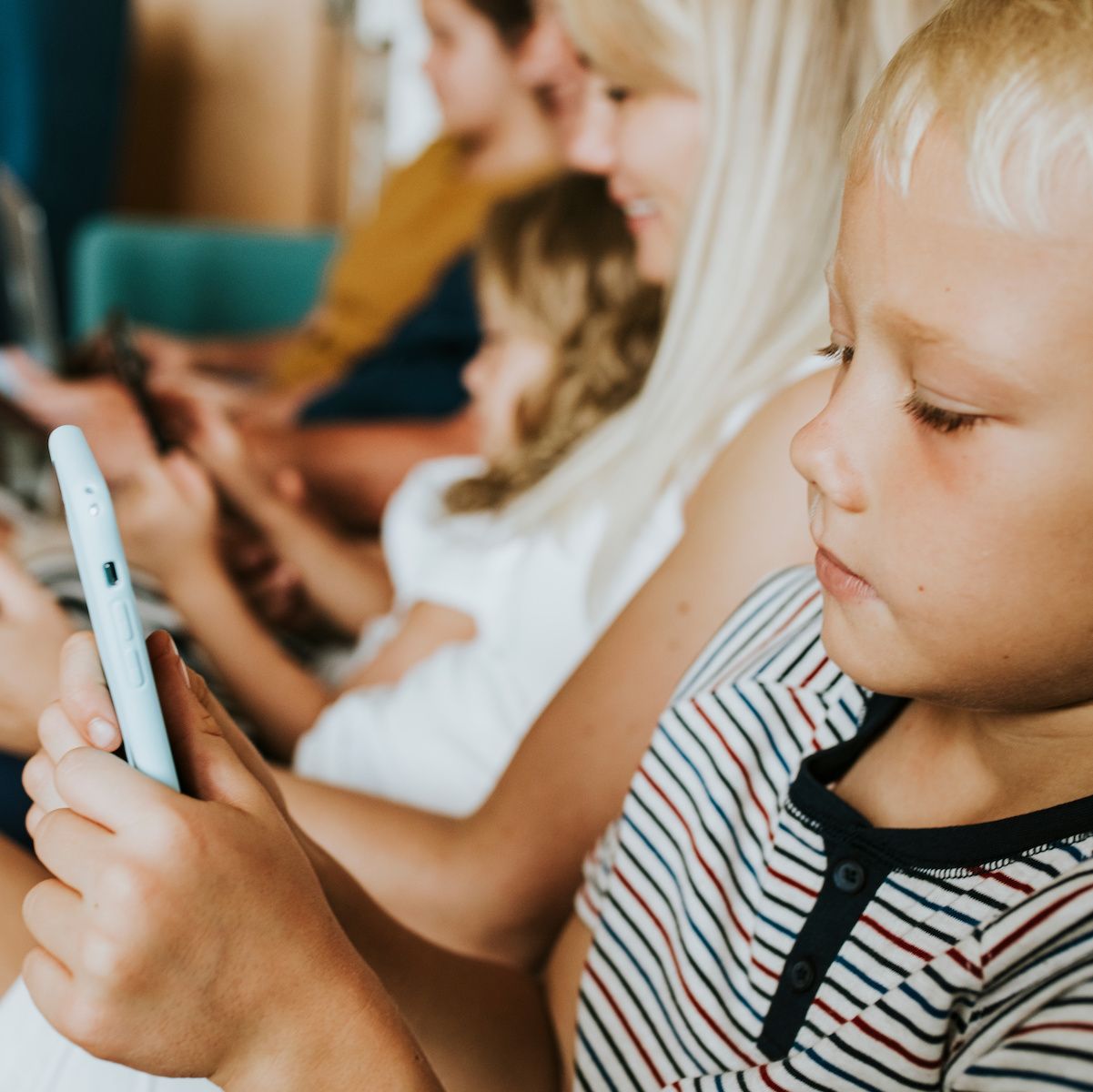 A young boy with blond hair sits on the couch with his siblings and uses internet data while playing on a cell phone.