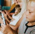 A young boy with blond hair sits on the couch with his siblings and uses internet data while playing on a cell phone.