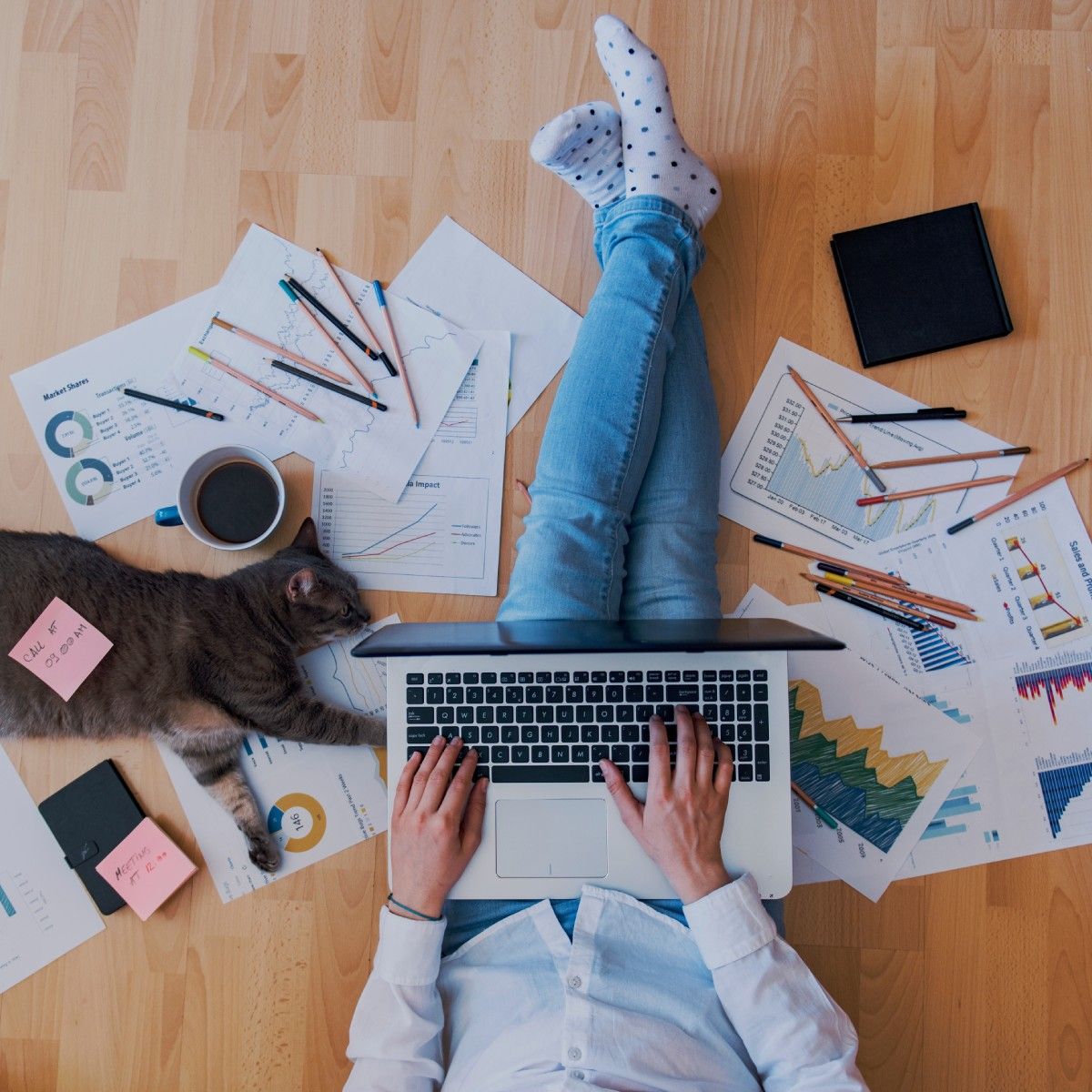 A top-down view of a person working from home, laptop in their lap and paper and pencils scattered around. Their cat is stretched out beside them.