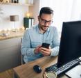 A white man with a beard and glasses sits in his home office and uses his desktop computer and cell phone