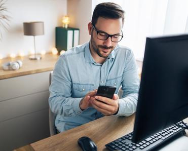 A white man with a beard and glasses sits in his home office and uses his desktop computer and cell phone