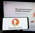 A hand holds a cell phone with the DuckDuckGo logo on the screen in front of the DuckDuckGo webpage.