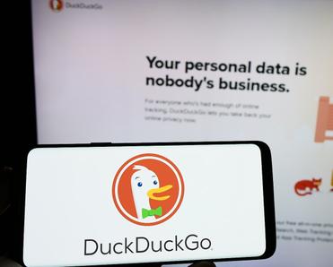 A hand holds a cell phone with the DuckDuckGo logo on the screen in front of the DuckDuckGo webpage.