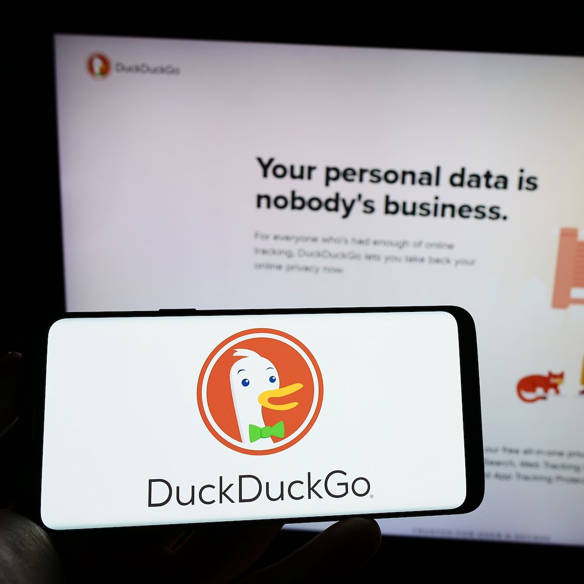 A hand holds a cell phone with the DuckDuckGo logo on the screen in front of the DuckDuckGo webpage.