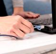 A close-up of a white woman&#x27;s right hand plugging a blue Ethernet cable into the side of her black laptop.