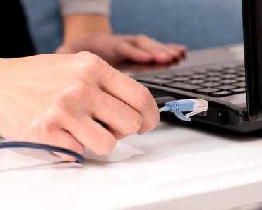 A close-up of a white woman&#x27;s right hand plugging a blue Ethernet cable into the side of her black laptop.